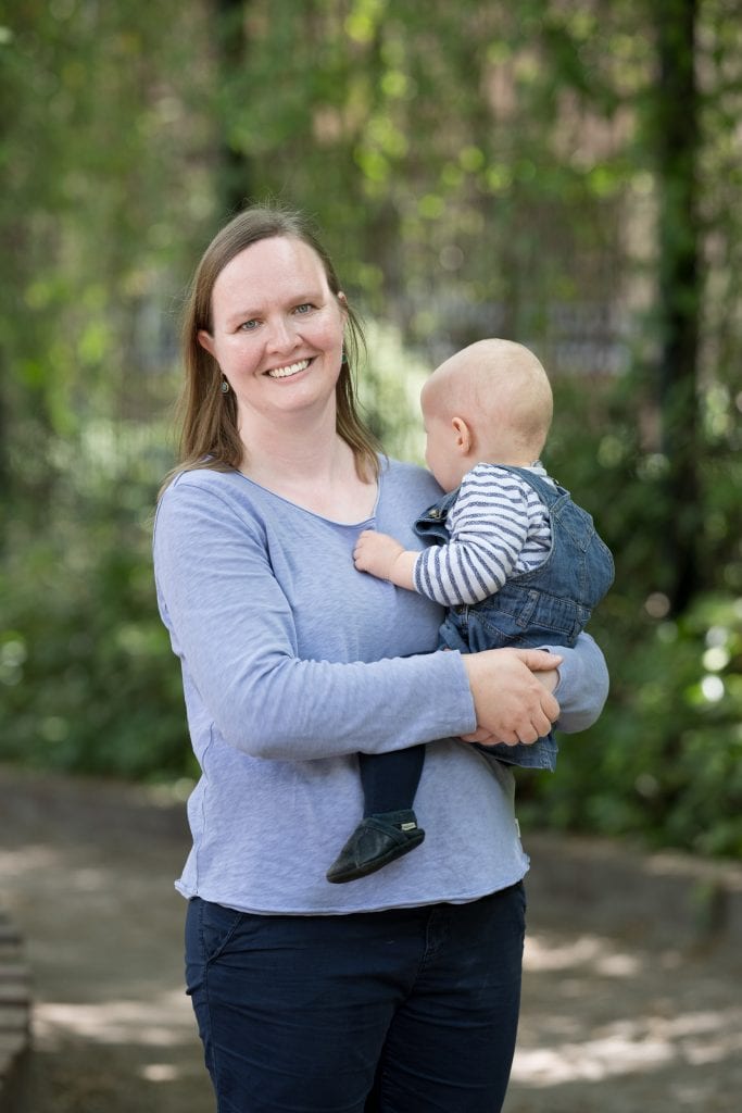 Miriam Stute steht draußen vor einem verschwommenen Hintergrund, der Pflanzen und einen grauen Boden vermuten lässt. Die Sonne scheint. Auf dem Arm hält sie ein Kleinkind, das das Kinn auf ihre linke Schulter legt und in den Bildhintergrund schaut. Miriam Stute blickt mit offenem Lächeln in die Kamera. Sie trägt ein langärmliges, hellblaues T-Shirt und eine dunkelblaue Hose. Ihre schulterlangen, glatten blonden Haare trägt sie offen.