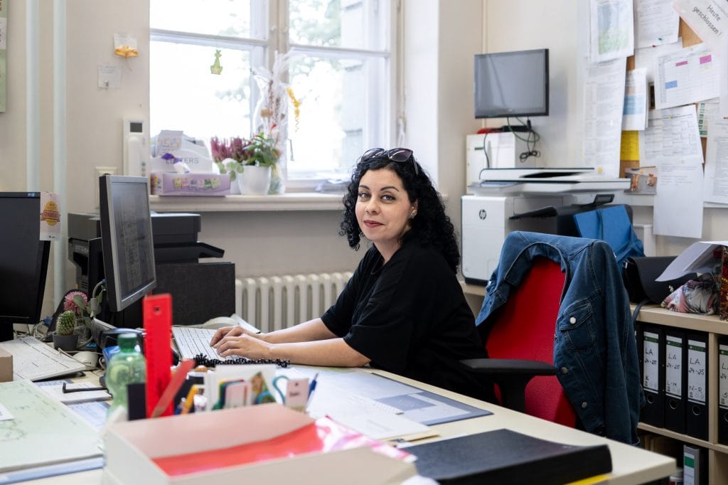 Ebru Schaefer sitzt auf einem Bürostuhl mit roter Lehne an einem großen Schreibtisch vor einem PC. Ihre Unterarme liegen auf dem Tisch ab, ihre Finger berühren eine Computertastatur, ihr Körper ist in Richtung der Tastatur und eines Bildschirms ausgerichtet, ihr Blick geht in Richtung Kamera, sie lächelt leicht. Auf dem Schreibtisch sind viele Büromaterialien, links im Bild ist ein weiterer Arbeitsplatz zu erkennen. Im Bildhintergrund ist eine Pinnwand voller bedruckter Zettel, ein kleines Regal, in dem Aktenordner stehen,  ist durch den Bürostuhl teils verdeckt. Auf dem Regal liegen eine schwarze Handtasche, ein blauer Stoffbeutel und ein Schal. Ebru Schäfer trägt ein schwarzes, weites, kurzärmliges Tshirt, ihre schwarzen, lockigen Haare liegen offen und gehen etwas über die Schulter, eine Sonnenbrille hält sie wie ein Haarreif aus dem Gesicht. An ihrem linken Ohr trägt sie einen  silbernen Ohrring und einen Piercing, der weiß glitzert. Ihre Augen sind geschminkt, sie trägt Lippenstift.