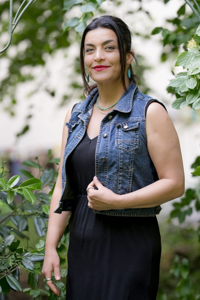 Eylem Bozkaya steht draußen vor einem lichten Strauch mit grünen Blättern und einigen weißen Blüten. Sie blickt mit einem Lächeln in die Kamera. Ihre dunkelbraunen, glatten Harre trägt sie in einem Zopf, links und rechts liegen Strähnen um ihr Gesicht. Ihre Augen sind leicht geschminkt, sie trägt roten Lippenstift. Sie trägt türkise Ohrringe und zwei Ketten, eine davon aus türkisenen Steinen. Über einem schwarzen, ärmellosen Kleid trägt sie offen eine ärmellose Jeansjacke. Ihre rechte Schulter dreht leicht in den Bildhintergrund, ihr rechter Arm hängt nach unten, während ihr linker Arm vor dem Oberkörper angewinkelt ist. An beiden Zeigefingern und an einem kleinen Finger trägt sie einen Ring.