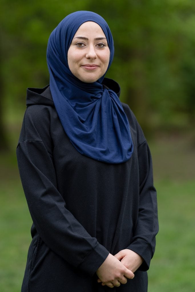 Esra Karamik sitzt draußen an einem Bistrotisch neben einer leicht befahrenen Straße. Auf der anderen Straßenseite reihen sich Läden aneinander. Esra Karamik ist links im Bild und blickt mit einem offenen Lächeln in Richtung einer zweiten Person rechts im Bild an der gegenüberliegenden Seite des Tisches. Beide haben ihre Ellenbogen auf dem Tisch liegen und sind zueinander gewandt. Esra Karamik stützt ihren Kopf auf ihre rechte Hand, am Handgelenk trägt sie einen goldenen Armreif. Sie trägt ein Nasenpiercing, ihre Augen sind leicht geschminkt. Beide Frauen tragen schwarze, langärmelige Oberteile und einen Hijab in dunkelblau bzw. hellbraun. Vor ihnen steht jeweils ein Kaffeebecher, daneben liegen ihre Mobiltelefone.