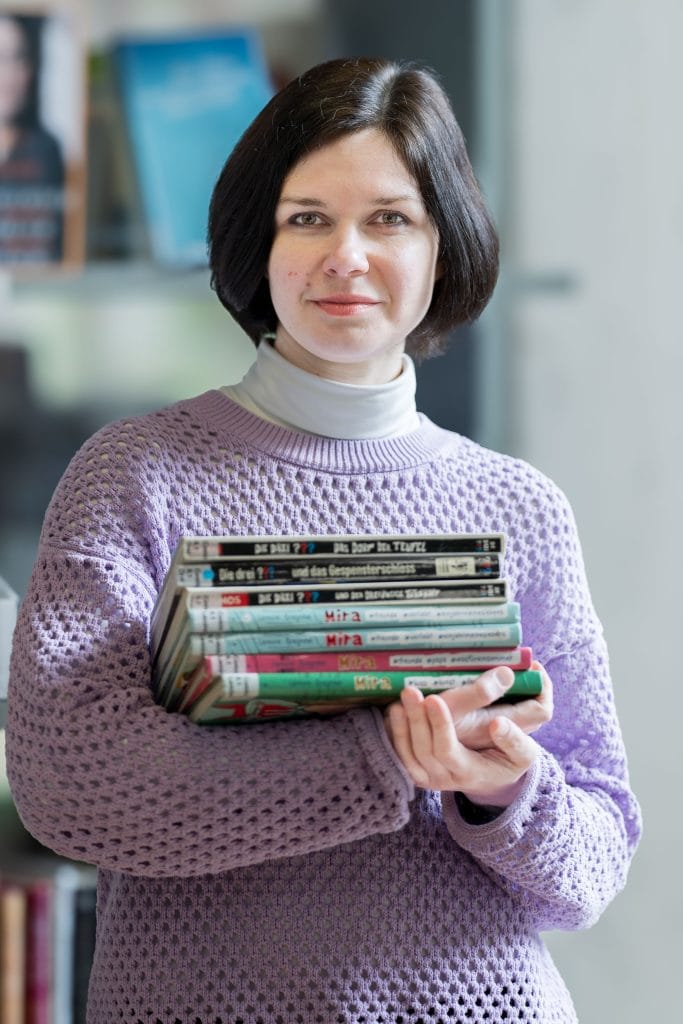 Nadjeschda Pugatschowa steht vor einem Bücherregal, das verschwommen zu erkennen ist. Sie blickt mit einem leichten Lächeln in die Kamera. Ihr rechter Arm ist vor ihrem Körper angewinkelt und wird von ihrem linken Arm gestützt. So hält sie einen Bücherstapel aus 3 Büchern von “Die drei Fragezeichen” und 4 Büchern aus einer Reihe mit dem Titel “Mira”. Die Bücher tragen Kennzeichnungen einer Bibliothek. Ihre glatten, dunkelbraunen Haare trägt sie in einem Kurzhaarschnitt bis zum Kinn, unter ihrem langärmligen, fliederfarbenen Pullover trägt sie ein weißes Rollkragenshirt.