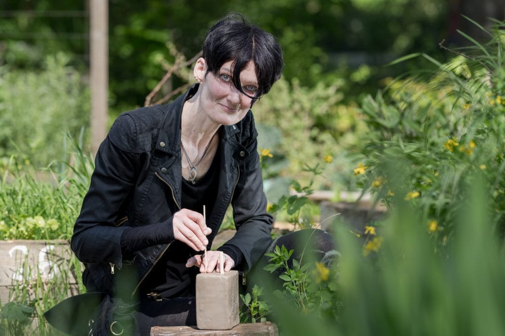 Jeanine Fornaçon kniet draußen zwischen Blumen und Gräsern, die teils in Kisten wachsen. Vor ihr liegt auf einem Holzblock ein Klumpen Ton, den sie mit zwei kleinen Holzstäben bearbeitet. Sie blickt mit einem Lächeln in die Kamera, eine schwarze Haarsträhne fällt aus ihrem Kurzhaarschnitt ins Gesicht. Über einem schwarzen Tshirt trägt sie offen eine Bikerjacke aus schwarzem Stoff. Sie trägt eine schwarze kurze Hose und schwarze Leggings. Um ihren Hals trägt sie an einem dünnen Lederband einen silbernen Anhänger